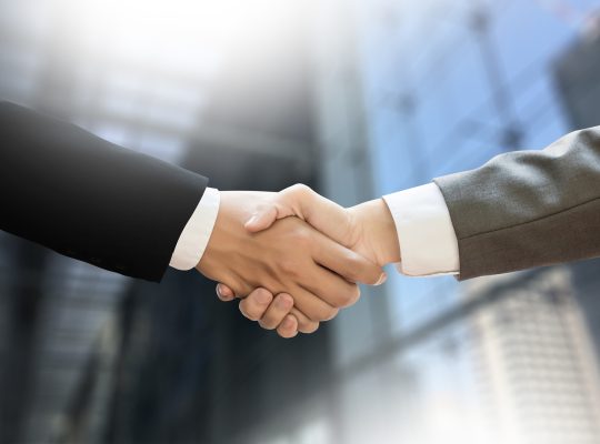 Dwóch biznesmenów w garniturach ściskających dłonie na tle zatłoczonej sali konferencyjnej